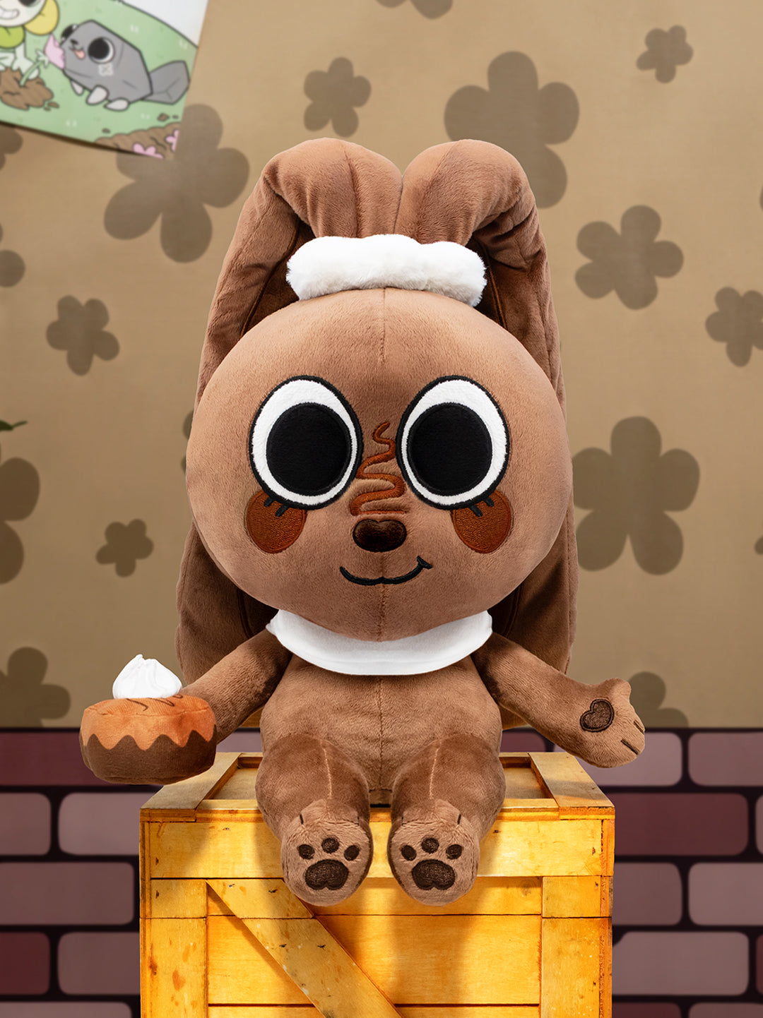 Plush toy resembling a cartoon character with a brown body and white accents, sitting on a wooden crate against a patterned wall.