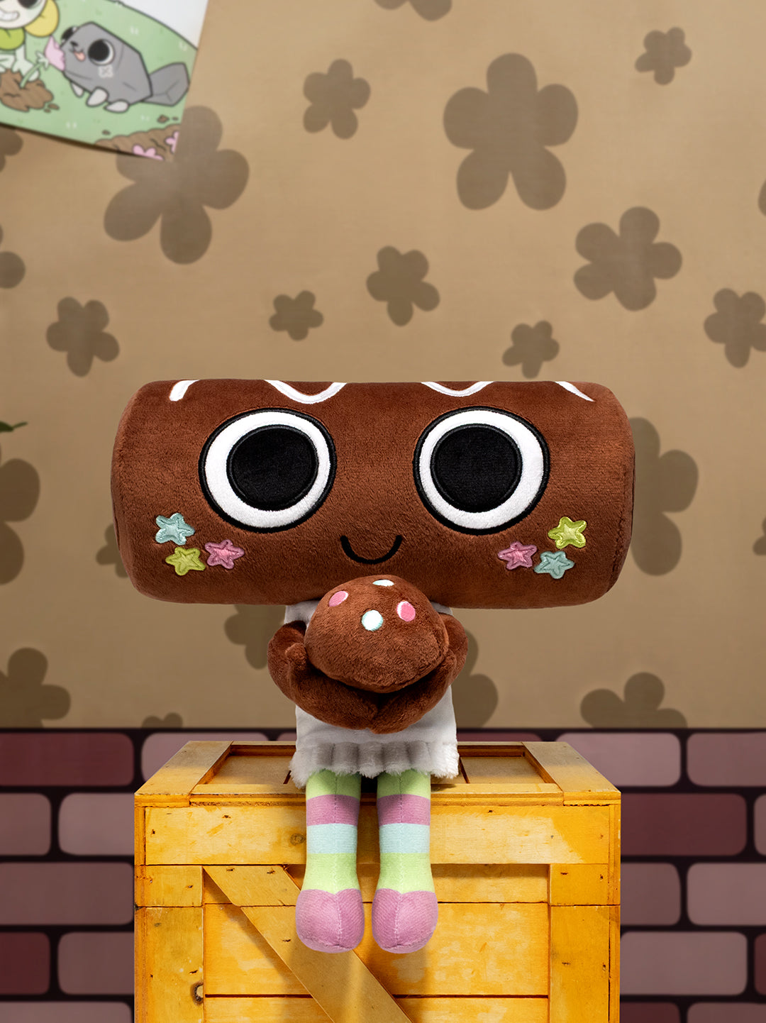 A plush toy shaped like a brown cookie or log with oversized black-and-white eyes, a smiling face, and small colorful star accents, sitting on top of a yellow wooden crate against a patterned tan wall.