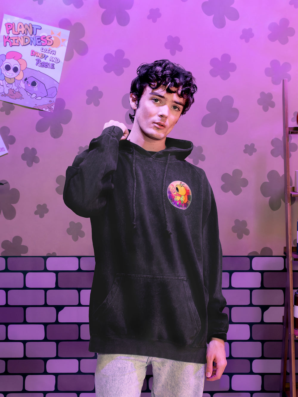 Person wearing a black hoodie with a colorful graphic design against a purple brick wall with flower patterns.