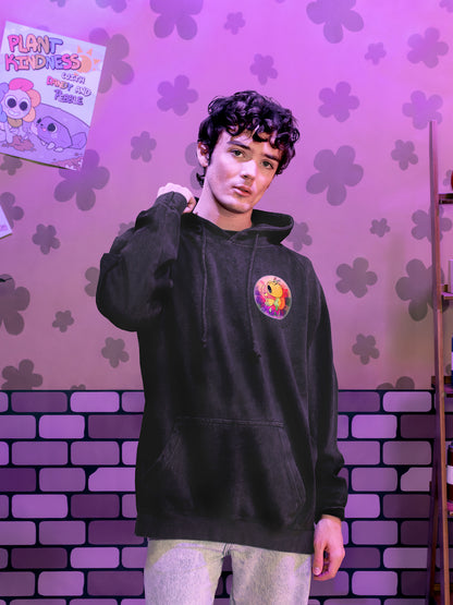 Person wearing a black hoodie with a colorful graphic design against a purple brick wall with flower patterns.