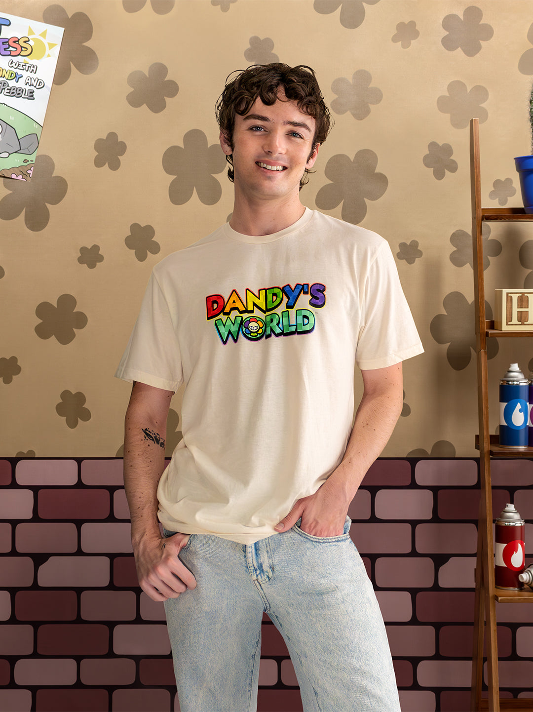 Person wearing a biege 'Dandy's World' t-shirt in front of a decorative wall with a shelf.
