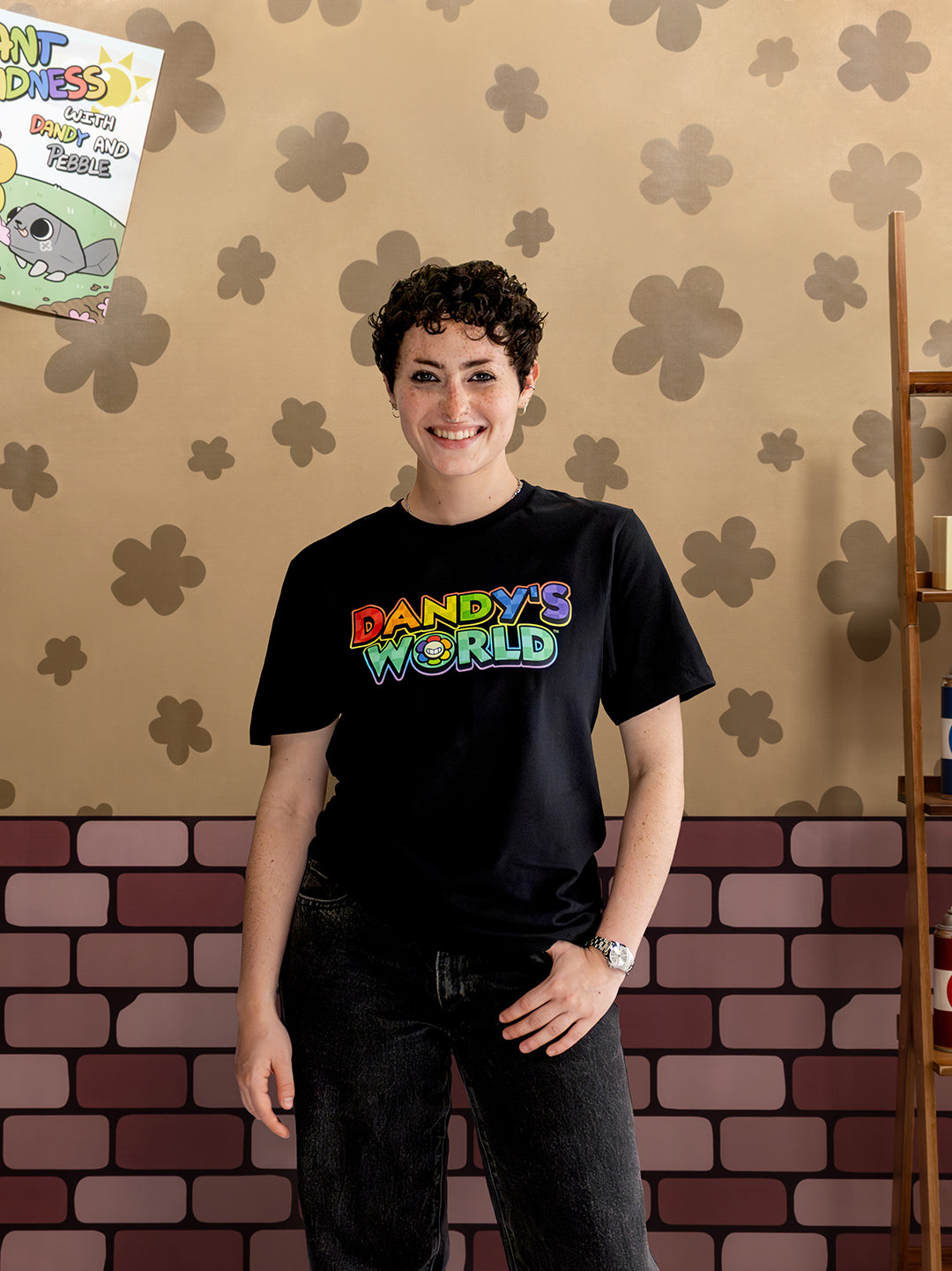 Person wearing a black t-shirt with 'Dandy's World' text, standing against a patterned wall.