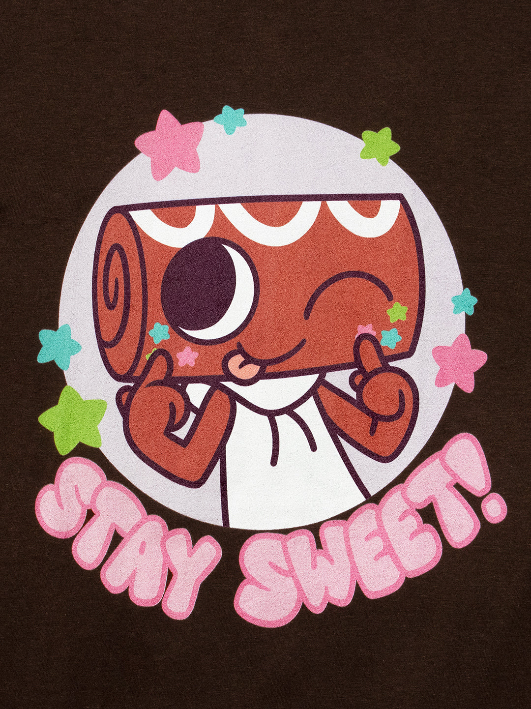 A close-up of a dark brown garment featuring a colorful cartoon graphic of a winking sweet roll character inside a white circle, surrounded by pink and green stars, with the text 'STAY SWEET!' in pink bubble letters below.