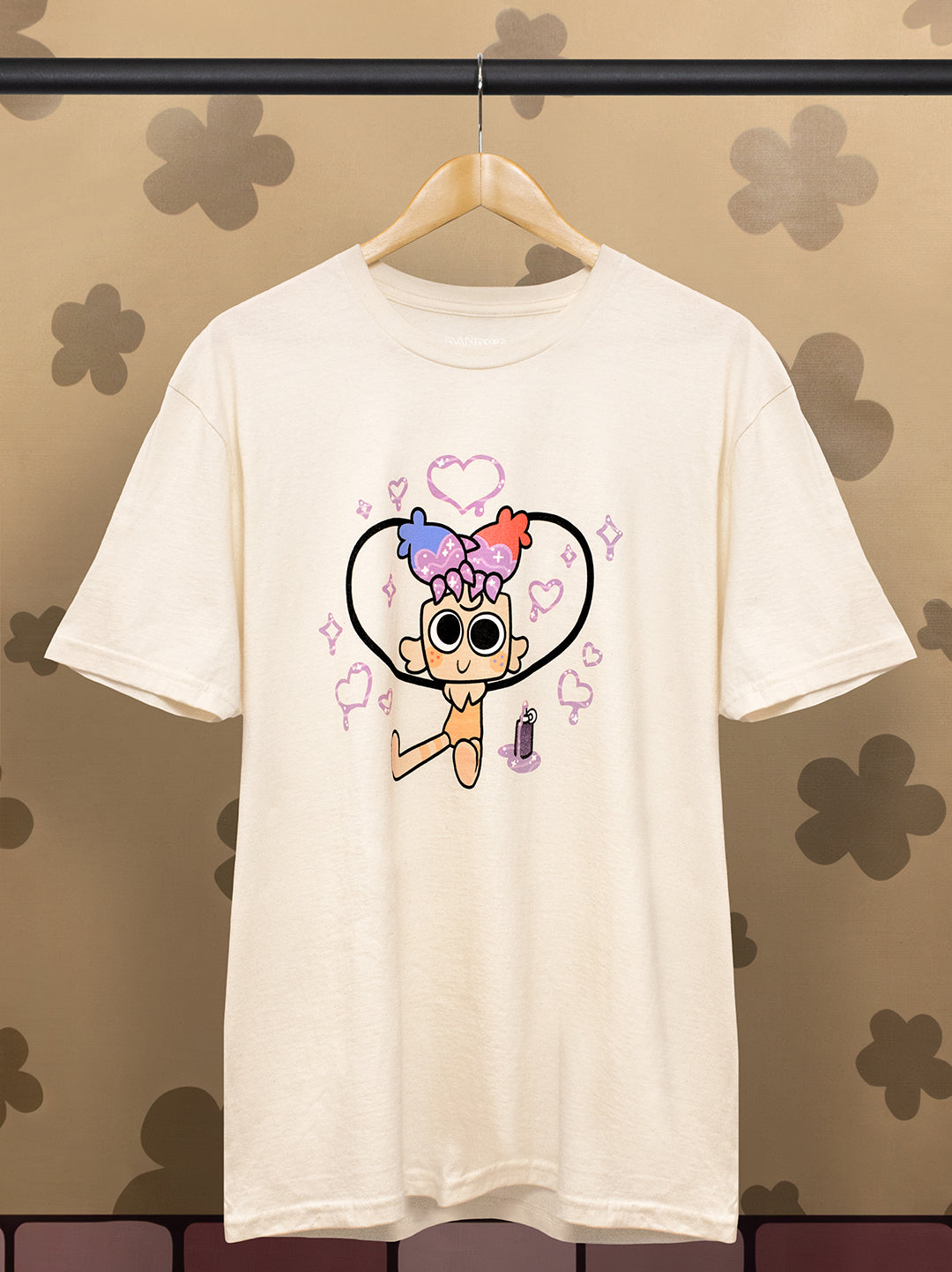 An off-white short-sleeve t-shirt hanging on a wooden hanger against a patterned brown wall. The shirt features a cartoon graphic of a character with colorful hair and sunglasses inside a heart outline.