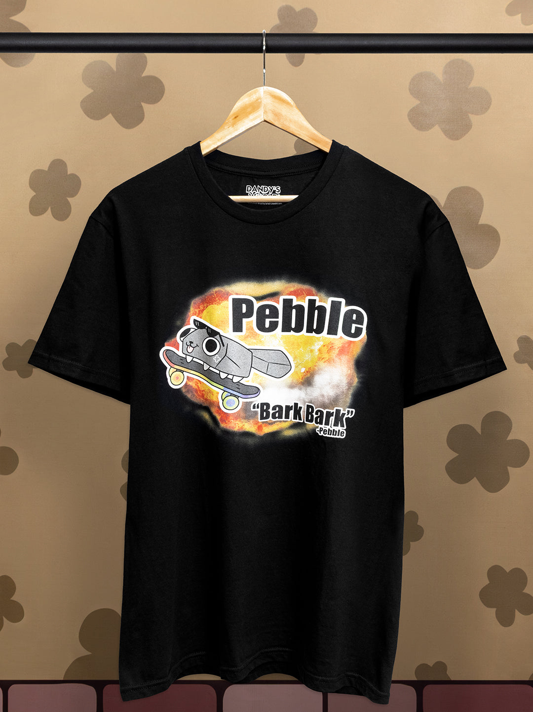 Black t-shirt with pebble riding a skateboard with explosions. text says "bark bark" - pebble. on a hanger against a patterned wall.