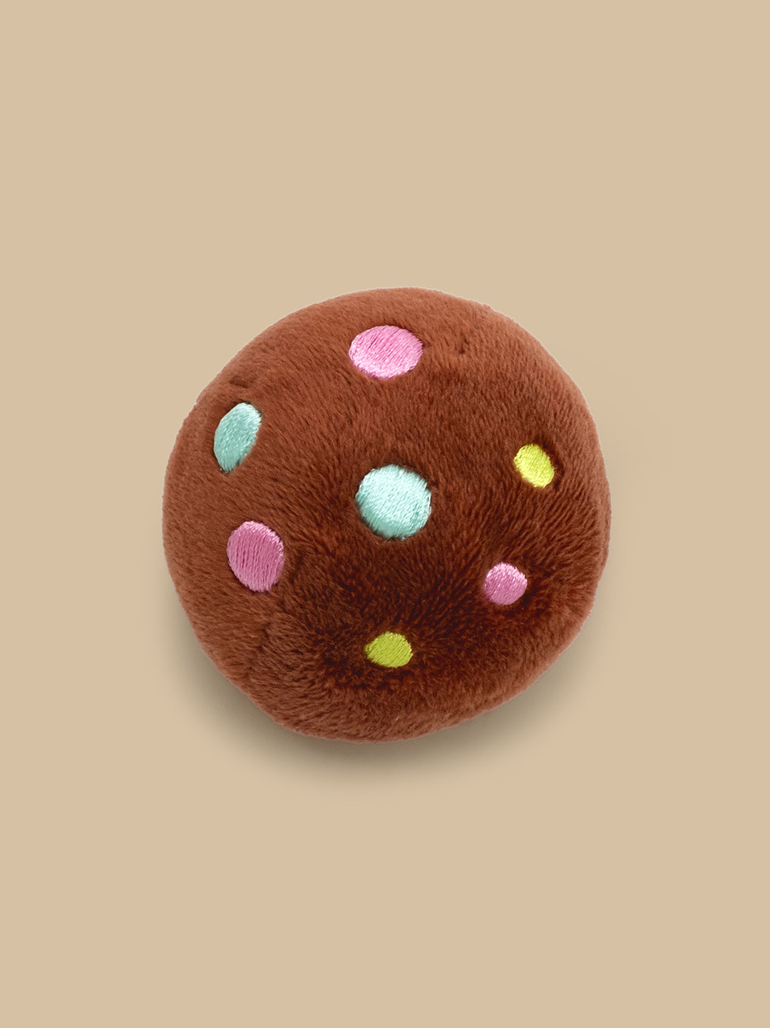 A plush round brown cookie toy with embroidered pink, light blue, and yellow dots, shown on a solid beige background.