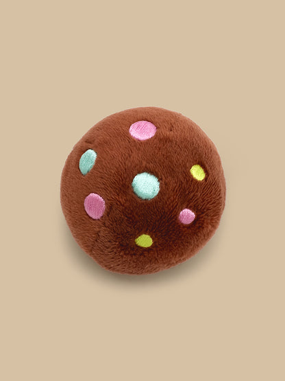 A plush round brown cookie toy with embroidered pink, light blue, and yellow dots, shown on a solid beige background.