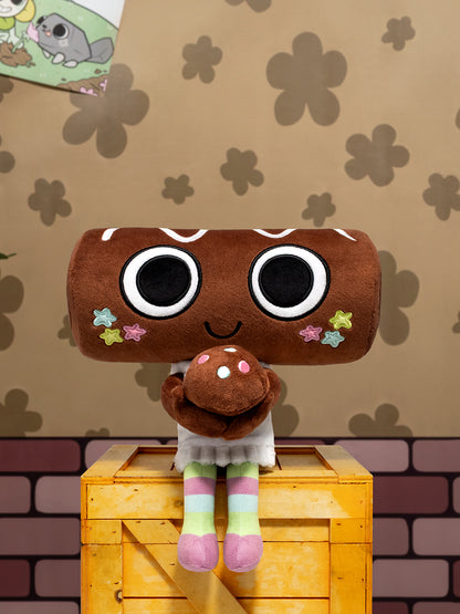 A plush toy shaped like a brown cookie or log with oversized black-and-white eyes, a smiling face, and small colorful star accents, sitting on top of a yellow wooden crate against a patterned tan wall.