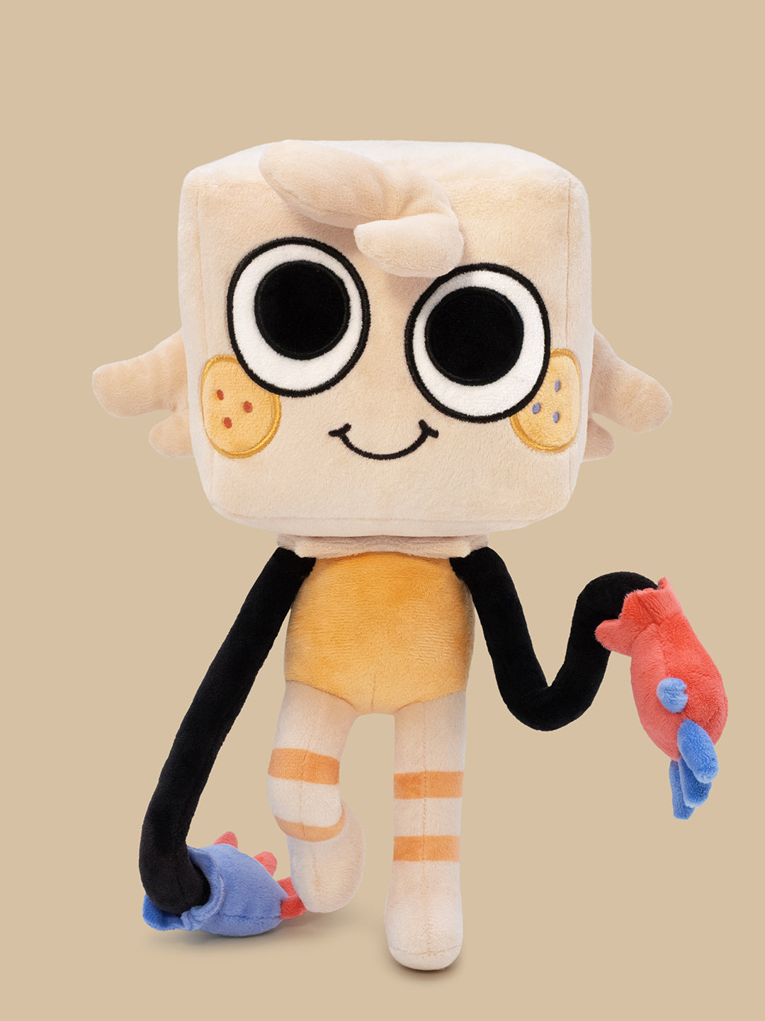 A plush toy character with a square cream head, large black-and-white eyes, a small smile, black arms, orange shorts, and colorful hands in red, blue, and light blue, shown against a plain beige background.