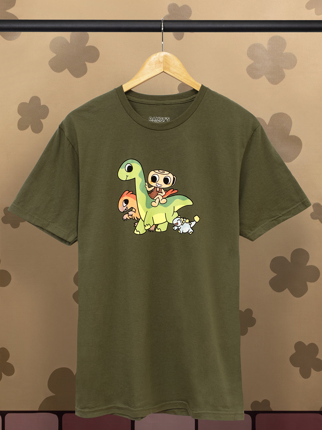 Green t-shirt with cartoon dinosaur design on a hanger against a floral-patterned wall.