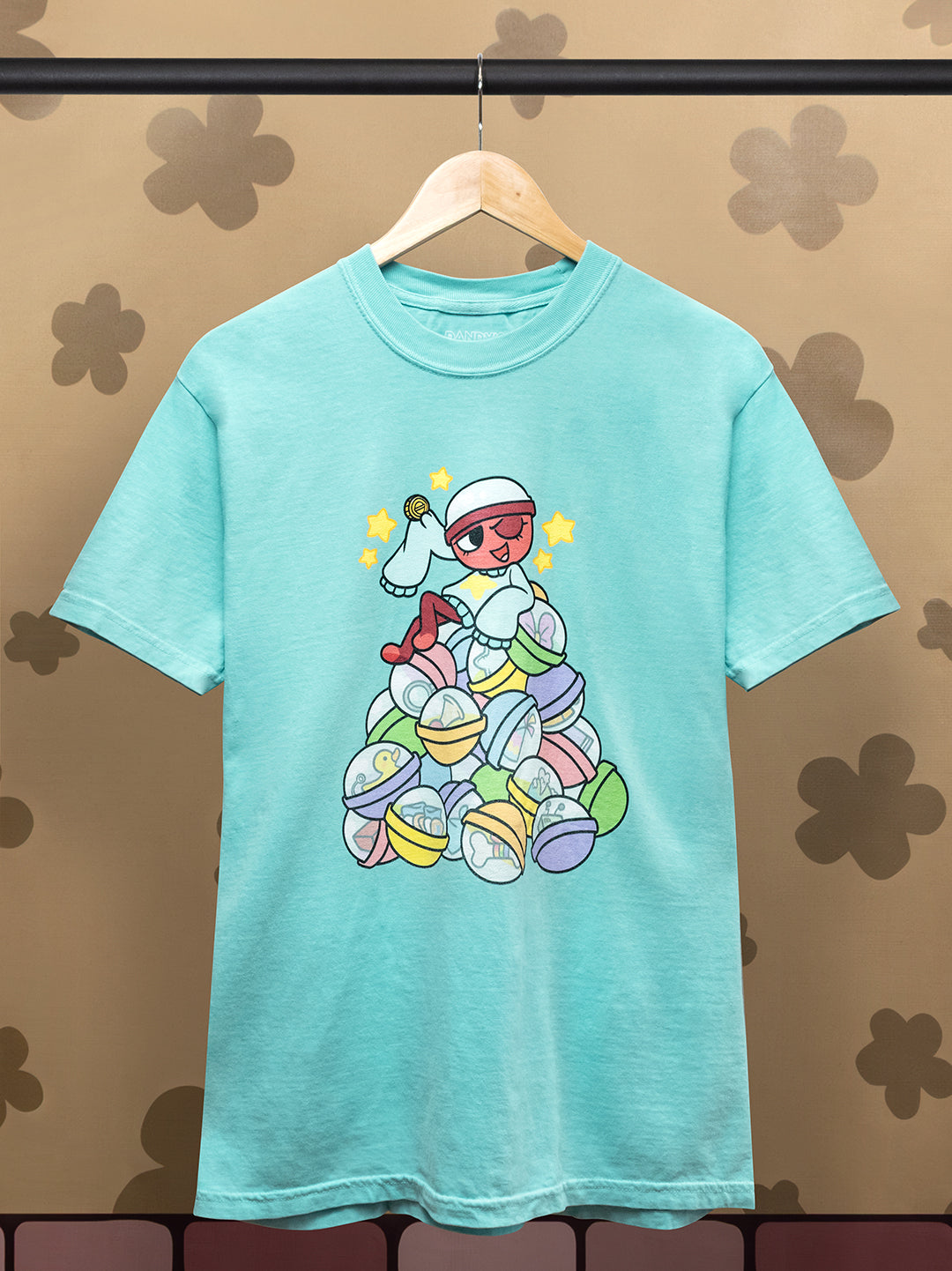 light blue t-shirt on a hanger. graphic: gigi sitting on top of gachapon balls with items and trinkets inside. Holding her lucky coin with stars around her head. She's winking. 