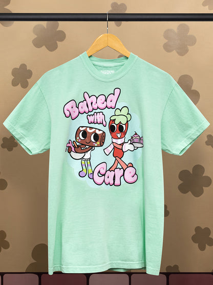 Mint green t-shirt with 2 cartoon characters and text saying "Baked with Care" on a hanger against a patterned wall.